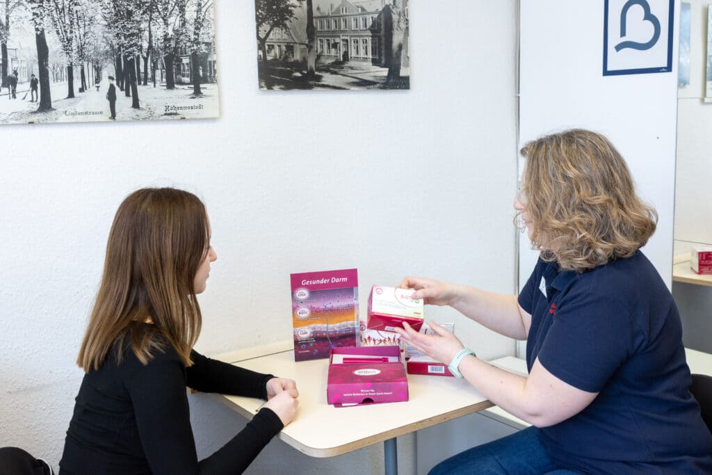 Zwei Frauen sitzen an einem Tisch mit Packungen von Gesundheitsprodukten und diskutieren über Vorsorge, während die eine der anderen eine Packung erklärt. An der Wand hinter ihnen hängen Schwarz-Weiß-Fotos.